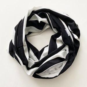 May include: Black and white striped infinity scarf. The scarf is made of a soft fabric and features a repeating pattern of wide black and white stripes. The scarf is looped and has a hidden pocket. The label reads "Robin's Dandies".