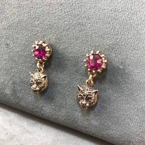 Puede incluir: Un par de pendientes de tono dorado con un cristal rosa y un dije de cabeza de tigre.