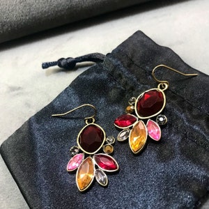 Op de afbeelding: Een paar gouden oorbellen met rode, oranje en roze edelsteenaccenten. De oorbellen hebben een grote ovale rode edelsteen in het midden, omringd door kleinere edelstenen in verschillende vormen en maten.