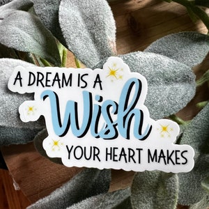 May include: A white sticker with the text "A dream is a Wish your heart makes" in black and light blue. The word "Wish" is in a cursive font. The sticker has yellow star accents and is set against a background of green leaves.