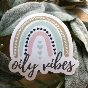May include: A sticker with a rainbow design in shades of green, pink, and brown. The rainbow has a white background with black hearts and a gold dotted border. The text "oily vibes" is written in black cursive below the rainbow.