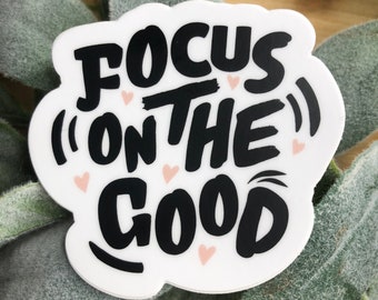 May include: White sticker with black text that reads "Focus (on the) good" with pink hearts scattered around the text.