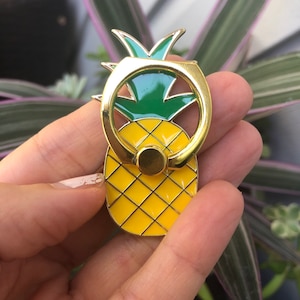 May include: A gold pineapple-shaped phone ring holder. The ring is attached to the top of the pineapple and is designed to be used as a stand for a phone.