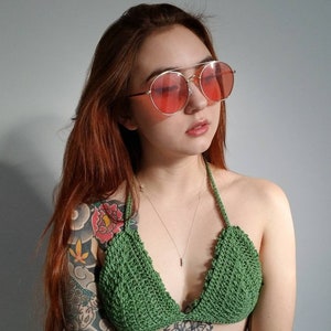 Könnte beinhalten: Eine Frau mit langen roten Haaren trägt ein grünes gehäkeltes Bikini-Oberteil mit einer silbernen Halskette. Sie trägt runde rosafarbene Sonnenbrillen.