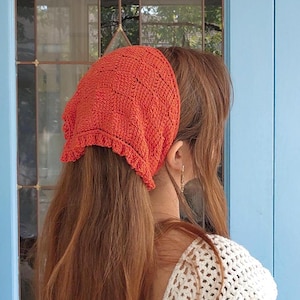 Puede incluir: Un pañuelo de ganchillo naranja con un borde con volantes. El pañuelo se lleva sobre un pelo largo y castaño rojizo. También se ve un top de ganchillo blanco.