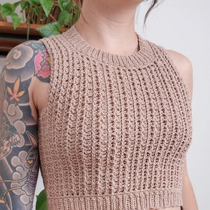 Op de afbeelding: Een bruin gebreid, mouwloos crop top met een geribbelde textuur. Het topje wordt gedragen met een bruine riem en een bruine broek.