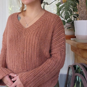 May include: A rust-coloured, ribbed knit jumper with a deep V-neck. The long-sleeved jumper has a relaxed fit and is made of a soft, textured material. The jumper is a warm, neutral colour, perfect for autumn or winter.