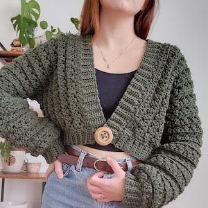 May include: A green crocheted cropped cardigan with a wooden button closure. The cardigan has a V-neck and long sleeves.