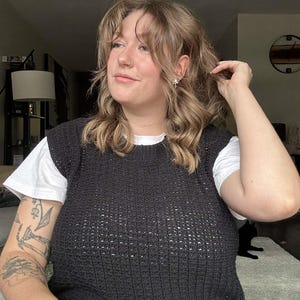 May include: A woman wearing a black crocheted sleeveless sweater over a white t-shirt. She has long, wavy brown hair and is wearing a nose ring.