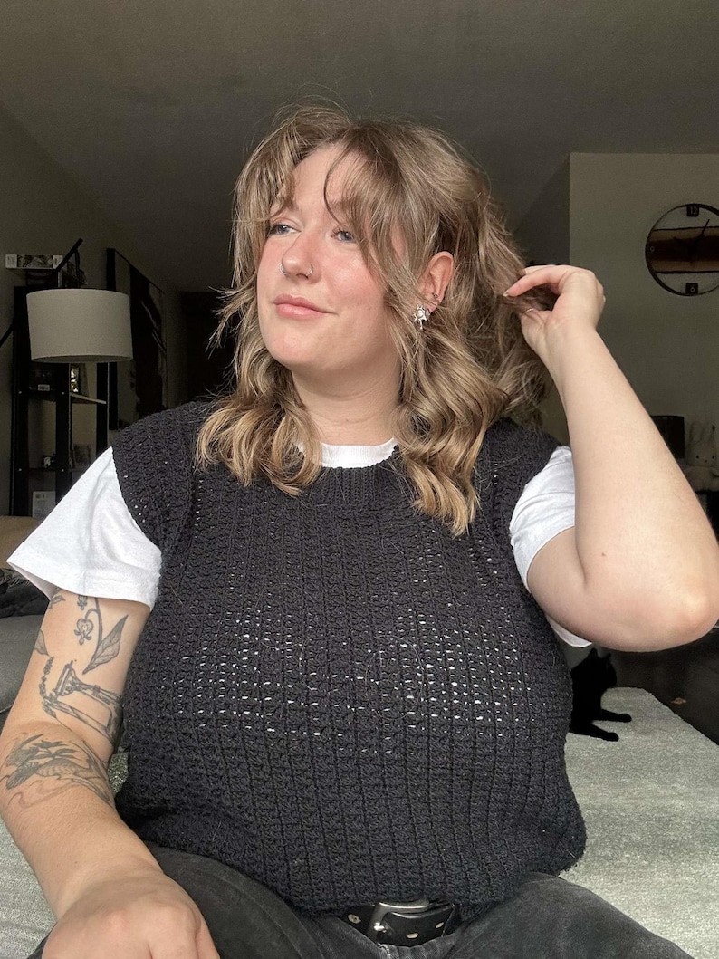 May include: A woman wearing a black crocheted sleeveless sweater over a white t-shirt. She has long, wavy brown hair and is wearing a nose ring.