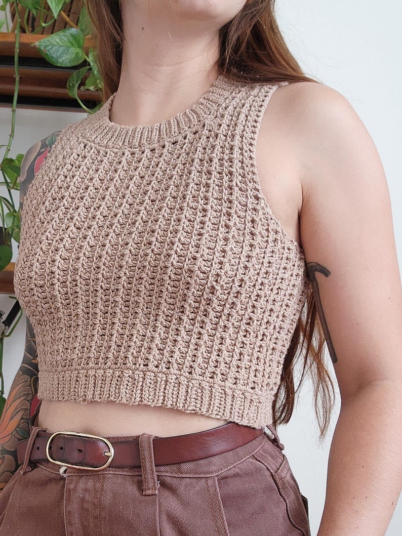 May include: A brown crocheted sleeveless crop top with a ribbed texture. The top is worn with a brown leather belt.