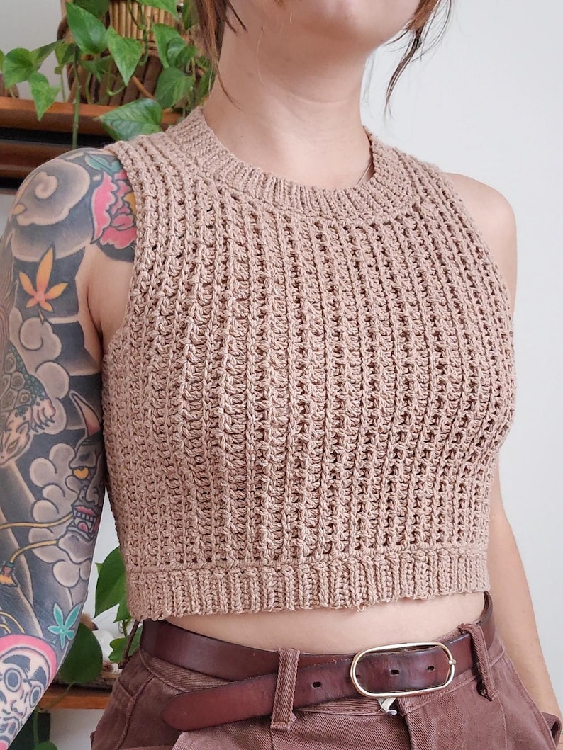 May include: A brown crocheted sleeveless crop top with a ribbed texture. The top is worn with a brown belt and brown trousers.