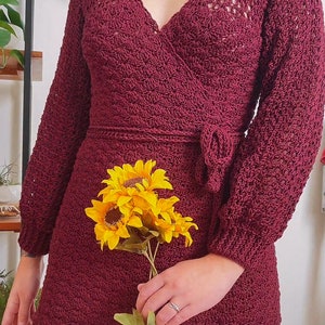May include: A maroon crocheted wrap dress with long sleeves and a ruffled hem. The dress has a V-neckline and a tie at the waist. The model is holding a bouquet of yellow sunflowers.