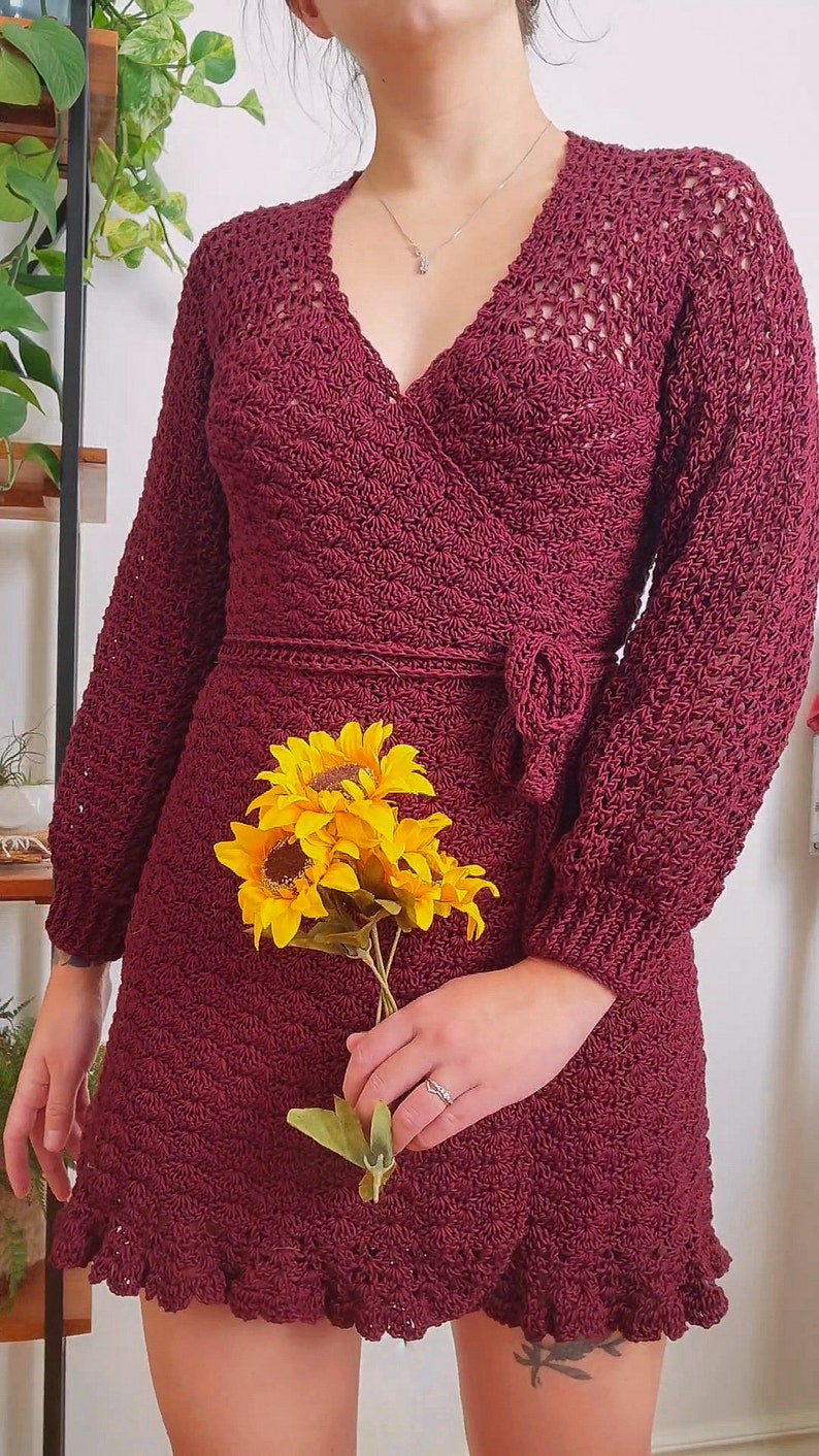 May include: A maroon crocheted wrap dress with long sleeves and a ruffled hem. The dress has a V-neckline and a tie at the waist. The model is holding a bouquet of yellow sunflowers.