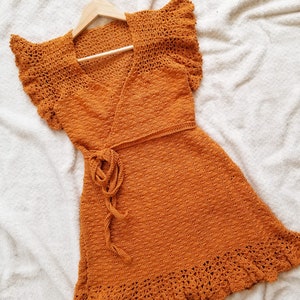 May include: A burnt orange crochet dress with a wrap style bodice and a ruffled hem. The dress is hanging on a wooden hanger.