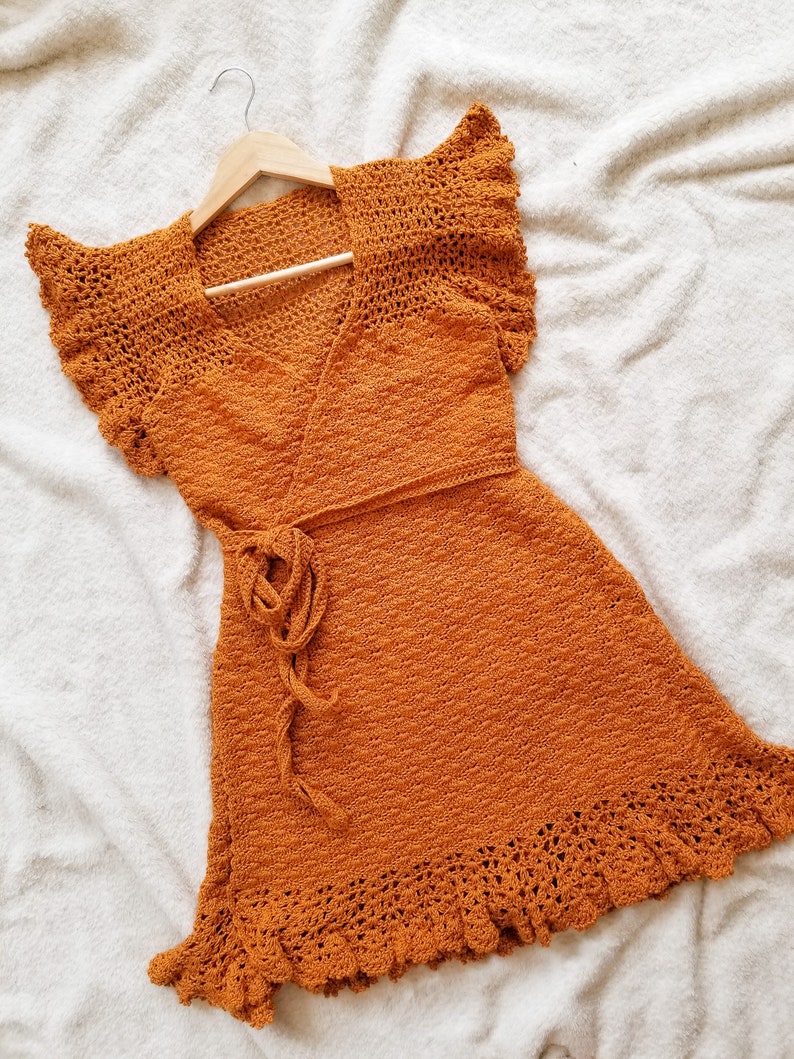 May include: A burnt orange crochet dress with a wrap style bodice and a ruffled hem. The dress is hanging on a wooden hanger.