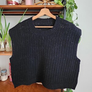 May include: A black sleeveless knitted sweater with a round neckline. The sweater is hanging on a wooden hanger.