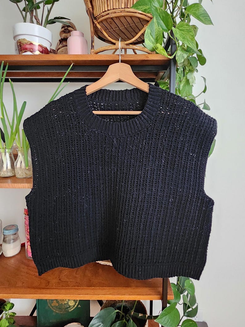 May include: A black sleeveless knitted sweater with a round neckline. The sweater is hanging on a wooden hanger.