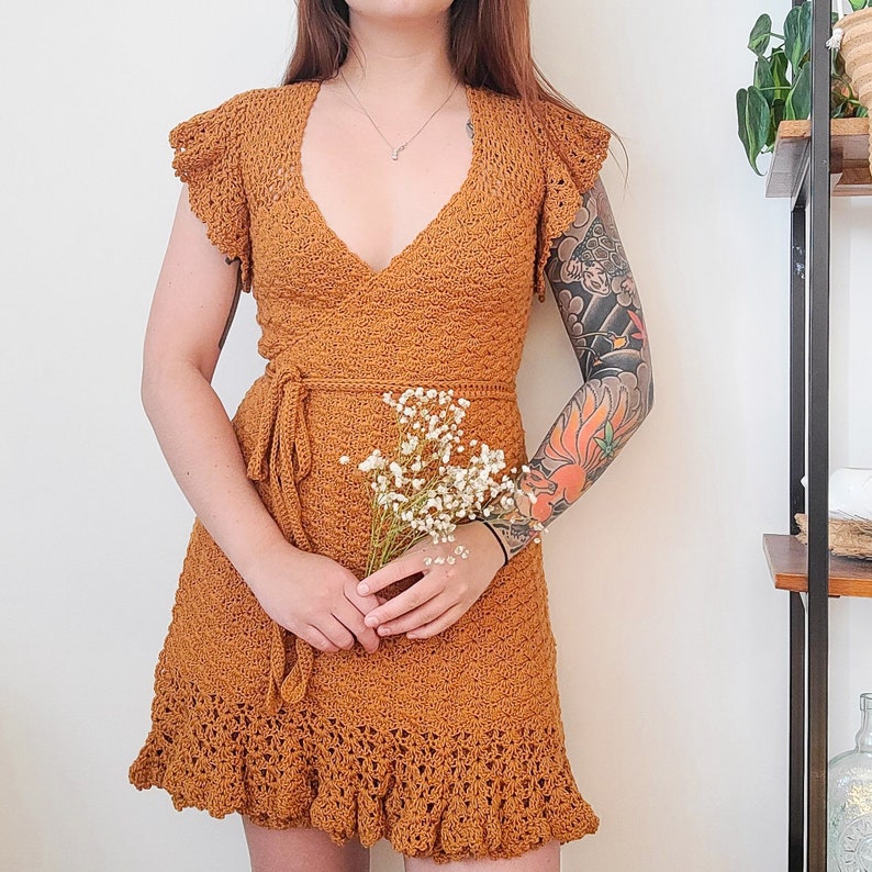 May include: A crocheted dress with a wrap-style bodice and a ruffled hem. The dress is a warm brown colour and is made of a delicate, open-weave fabric.