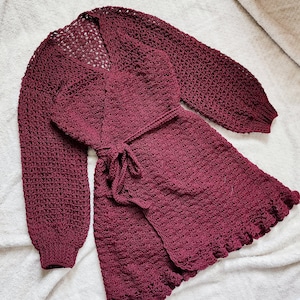 May include: A maroon crocheted cardigan with a wrap style and long sleeves. The cardigan has a tie belt and a ruffled hem.