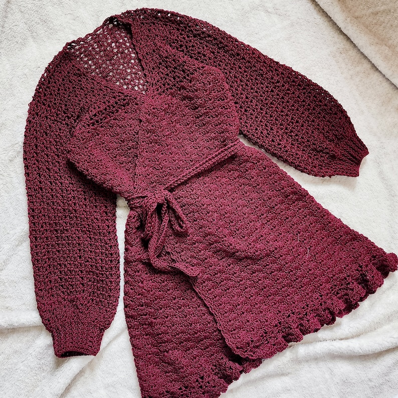 May include: A maroon crocheted cardigan with a wrap style and long sleeves. The cardigan has a tie belt and a ruffled hem.