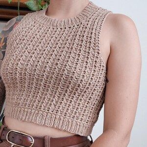 May include: A brown crochet tank top with a loose, open weave. The top is sleeveless and has a round neckline. It is cropped and hits just above the waist.