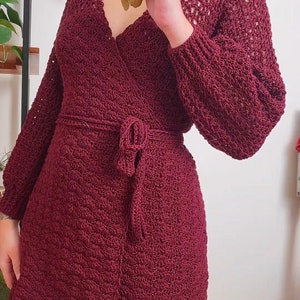 May include: A maroon crocheted wrap dress with long sleeves and a tie belt. The dress has a ruffled hem and a delicate openwork pattern.