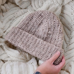 May include: A soft, light brown crocheted beanie hat with a ribbed texture. The hat is folded down and held in a hand.