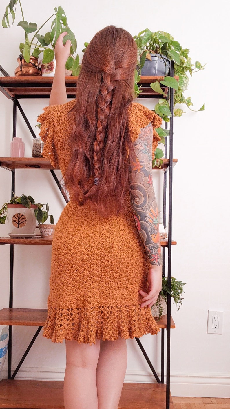 May include: A woman wearing a rust-coloured crochet dress with a braided hairstyle and a large tattoo on her arm. She is standing in front of a shelf with plants.