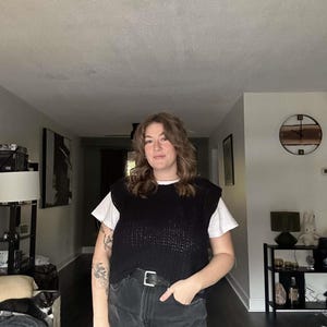 May include: A person wearing a black knitted vest over a white t-shirt, black jeans, and a black belt. The person has brown hair and is standing in a room with a clock on the wall.