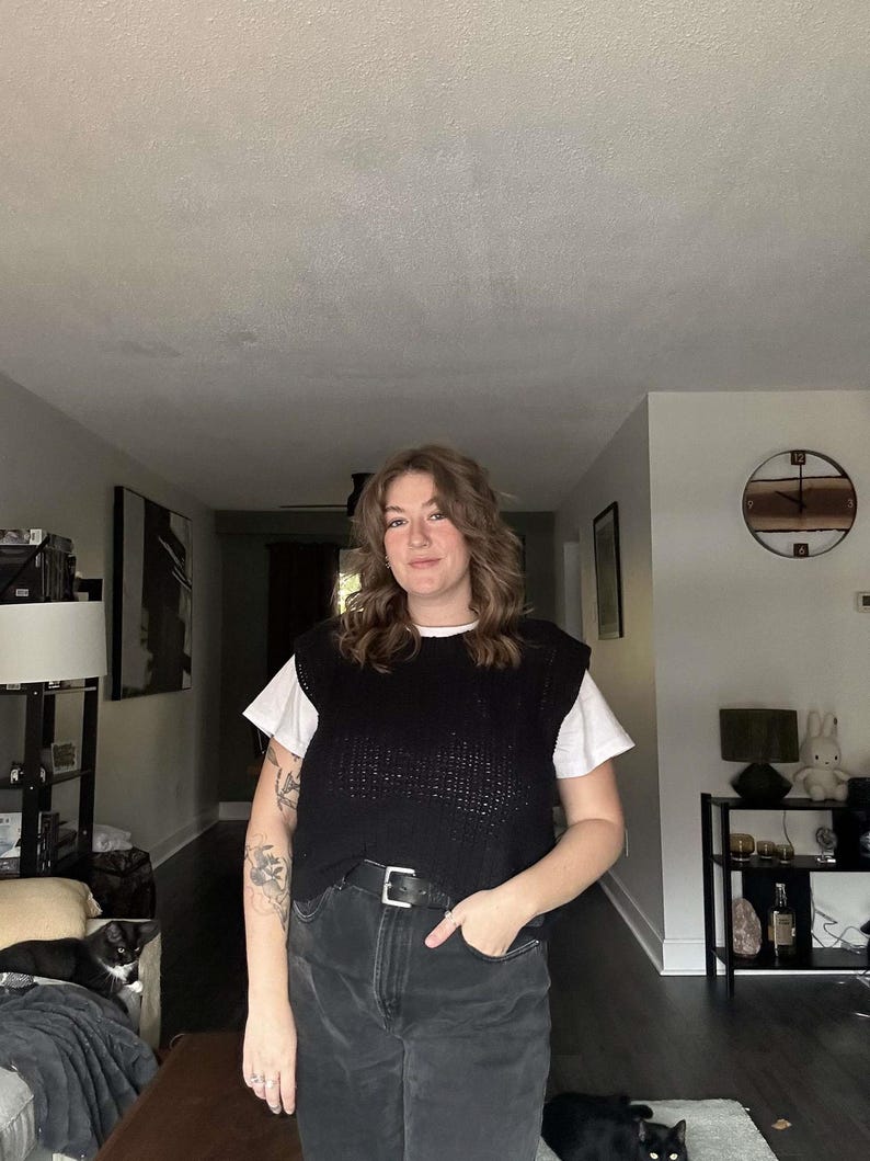 May include: A person wearing a black knitted vest over a white t-shirt, black jeans, and a black belt. The person has brown hair and is standing in a room with a clock on the wall.