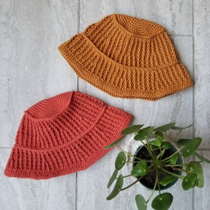 May include: Two crocheted bucket hats, one in a burnt orange color and the other in a coral color. The hats are sitting on a light gray tiled surface.