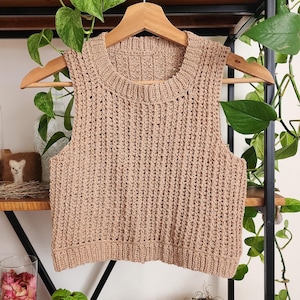 May include: A brown crocheted sleeveless top with a round neckline. The top is hanging on a wooden hanger against a wooden shelf with green plants.