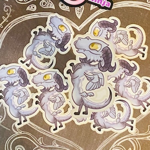 Pode incluir: Um conjunto de seis adesivos com ilustrações de desenhos animados de dragões cinzentos e brancos com olhos amarelos e asas. Os dragões estão em várias poses, alguns com as asas abertas e outros com as asas fechadas. Os adesivos estão sobre um fundo cinza escuro com desenhos florais brancos.