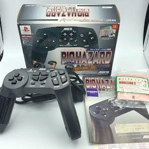 Puede incluir: Un mando negro de PlayStation con un cable gris, una caja de juego para el juego Biohazard y un folleto para Biohazard 2. La caja del juego tiene un diseño rojo y blanco con el texto "Biohazard" en letras blancas grandes. El folleto tiene un diseño rojo y blanco con el texto "Biohazard 2" en letras blancas grandes.