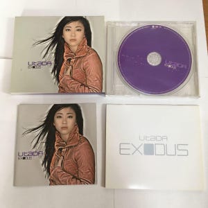 May include: CD album titled "Exodus" by Utada. The album cover features a woman with long black hair wearing a rust-colored jacket. The CD case is open, revealing a purple CD. The album title is also printed on the CD.