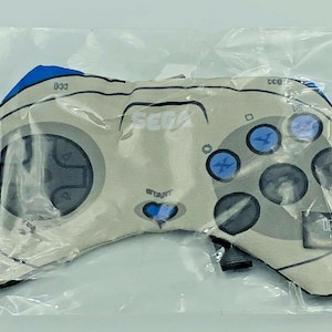 May include: A beige SEGA controller-shaped plush toy with blue accents. The controller has a directional pad, buttons, and the SEGA logo. The toy is packaged in clear plastic.