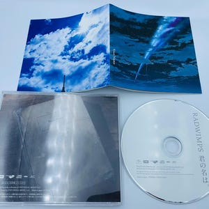 May include: A CD case with a silver CD inside. The front of the case features a blue sky with white clouds and a shooting star. The back of the case has text in Japanese and English. The text on the back of the case reads "RADWIMPS "Your Name" "
