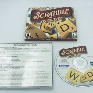 Może przedstawiać: Pudełko z grą komputerową i płyta CD do gry Scrabble Complete, gry słownej. Pudełko jest czerwone, białe i niebieskie z obrazkiem płytek Scrabble. Płyta CD jest srebrna z obrazkiem płytek Scrabble i napisem "Scrabble Complete".