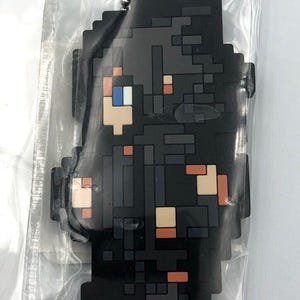 以下が含まれることがあります： 青い目をしたキャラクターが描かれた、黒とグレーのピクセルキーホルダー。キャラクターは赤とオレンジのシャツを着ています。