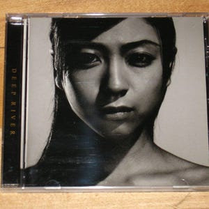 May include: A CD case featuring a black and white image of a woman's face. The woman has long dark hair and is looking directly at the viewer. The text "DEEP RIVER" is printed vertically on the left side of the case.