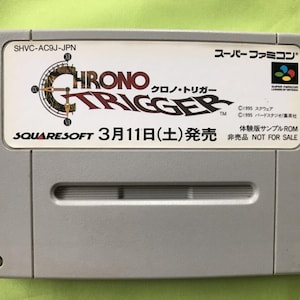 May include: A Super Nintendo Entertainment System (SNES) game cartridge for Chrono Trigger, a role-playing game. The cartridge is grey with a white label featuring the game's title, logo, and release date: 11 March 1995. The label also includes the game's copyright information and a note that the cartridge is a sample ROM and not for sale.