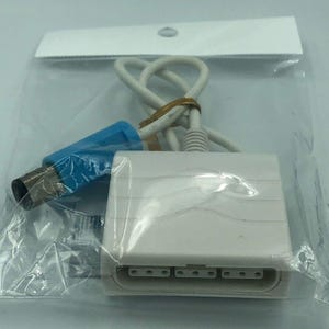 Könnte beinhalten: Ein weißer Videospielkonsolenadapter mit blauem Stecker und weißem Kabel, verpackt in einer transparenten Plastiktüte. Der Adapter hat mehrere Eingangsanschlüsse und ist mit einem Gummiband gesichert.