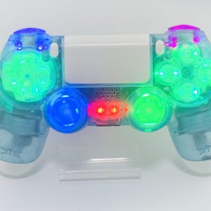May include: A transparent blue games controller with illuminated buttons and directional pads. The controller has green, blue, and red lights, with a white centre section. The design includes a clear stand.
