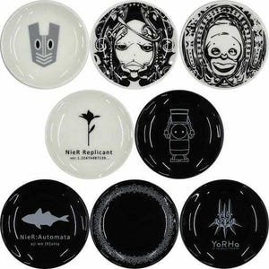 May include: A set of nine small, round ceramic plates. Each plate has a unique black and white graphic design, including stylized characters, floral motifs, and text. The plates are mainly white or black, with the designs in contrasting colours.