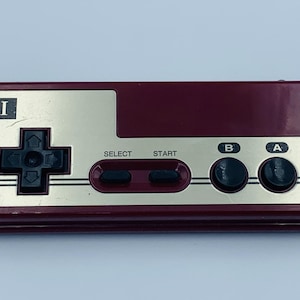 Könnte beinhalten: Ein weinroter und goldfarbener Retro-Videospiel-Controller. Der Controller hat ein Steuerkreuz, Select- und Start-Tasten sowie A- und B-Tasten. Der Buchstabe "I" ist in der oberen linken Ecke aufgedruckt.