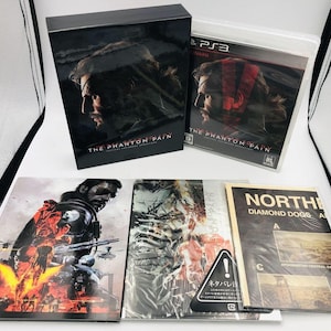 Può includere: Un set in edizione da collezione di Metal Gear Solid V: The Phantom Pain che include una scatola espositiva nera, una custodia per gioco PlayStation 3 e tre opuscoli illustrati. Gli articoli presentano illustrazioni e il titolo del gioco.