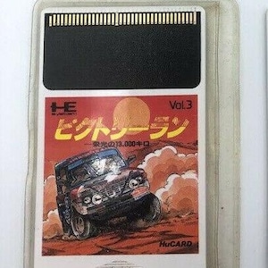 May include: A white plastic cartridge for the TurboGrafx-16 video game console. The cartridge has a black label with the text "Hucard" and a picture of a red jeep driving through a desert landscape. The text "Vol. 3" is in the top right corner of the label. The text "レーシング" is in the centre of the label. The text "光の213,000キロ" is below the picture.