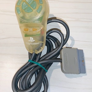 May include: A clear plastic PlayStation controller with a green button and a black cord. The controller has the word "PlayStation" printed on it.
