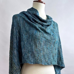 Può includere: Uno scialle tessuto blu e turchese con collo a imbuto, esposto su un manichino. Lo scialle presenta un motivo quadrato strutturato e pannelli lunghi e fluenti. La tavolozza dei colori comprende sfumature di turchese, blu e accenni di verde.